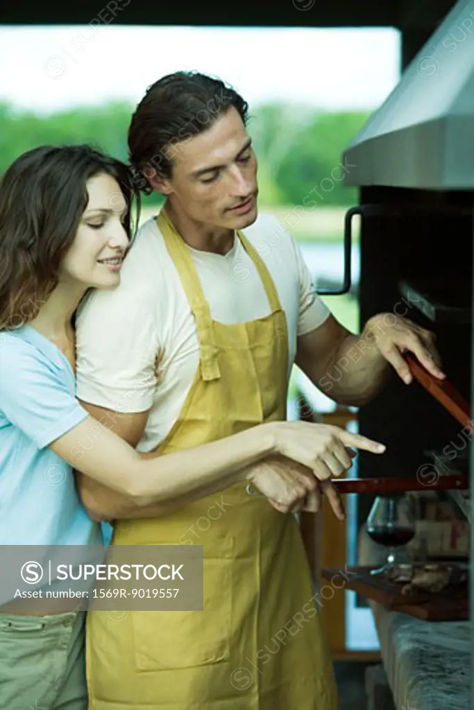 Man tending barbecue, while woman looks over his shoulder and points