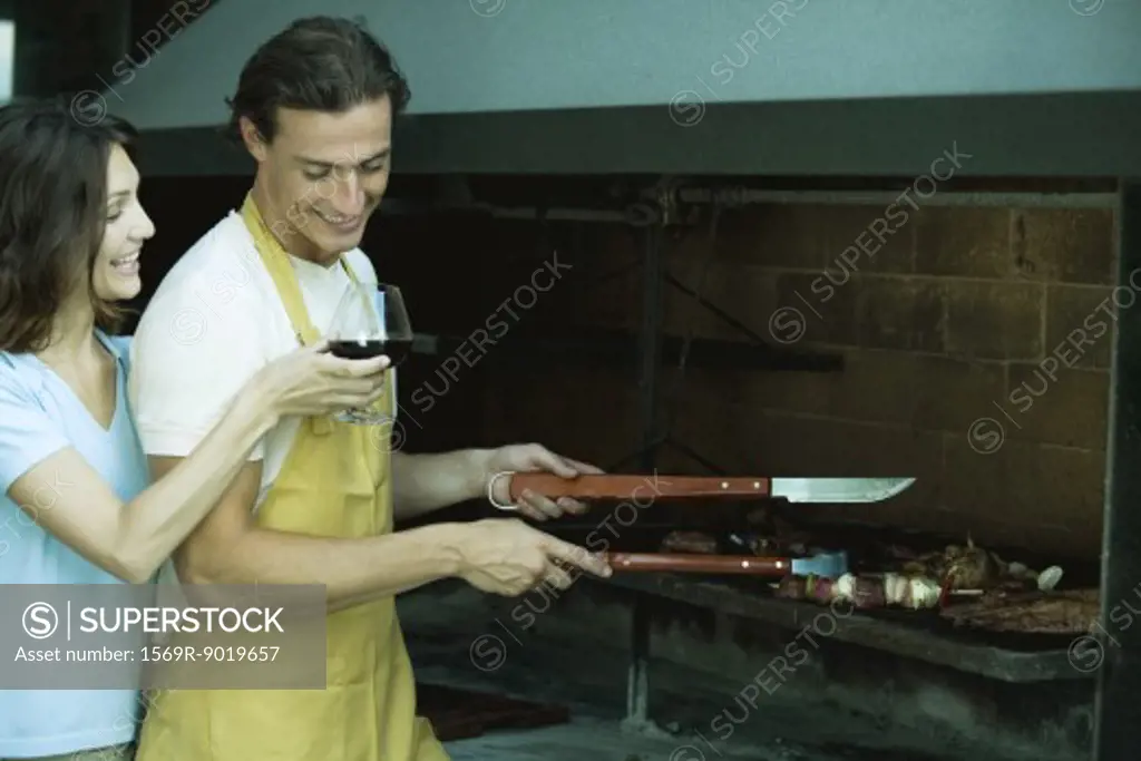 Man tending barbecue, while woman holds up glass of wine for him