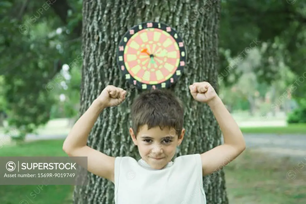 Boy standing in front of dart board on tree, arms up