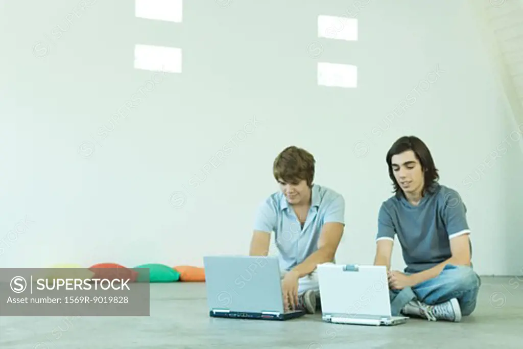 Two teen boys sitting on floor, using laptops