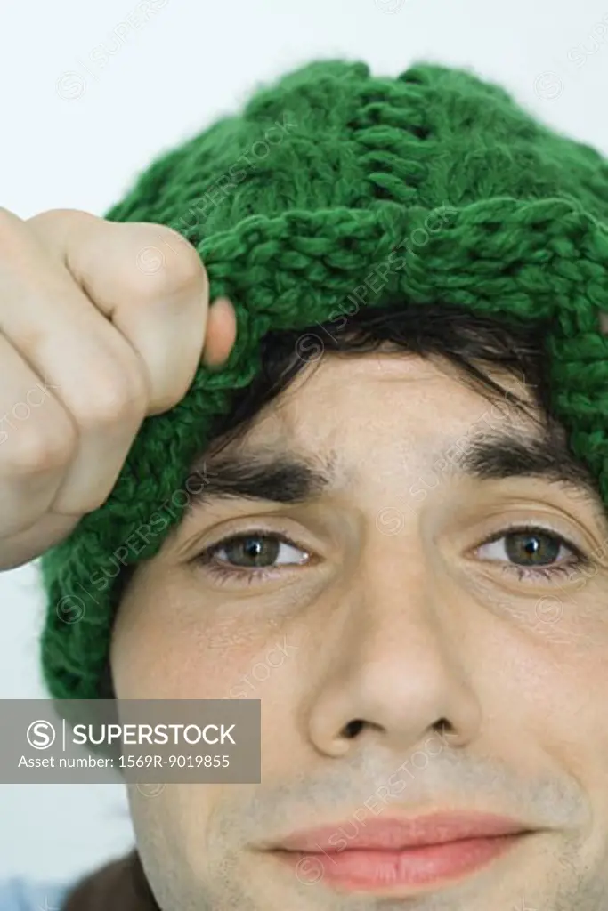 Young man lifting up edge of knit hat, looking at camera, close-up