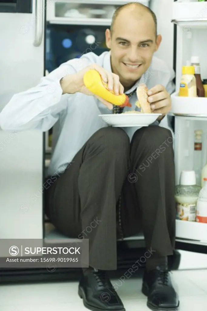 Man sitting in front of open refrigerator, putting mustard on hamburger