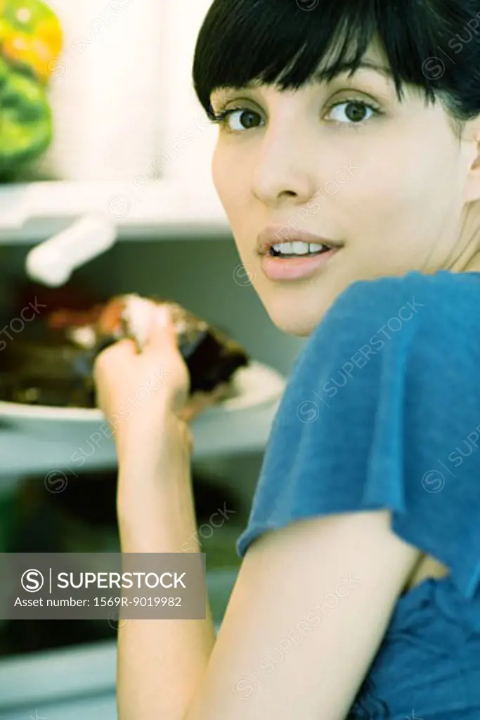 Woman taking piece of cake from refrigerator, looking over shoulder at camera