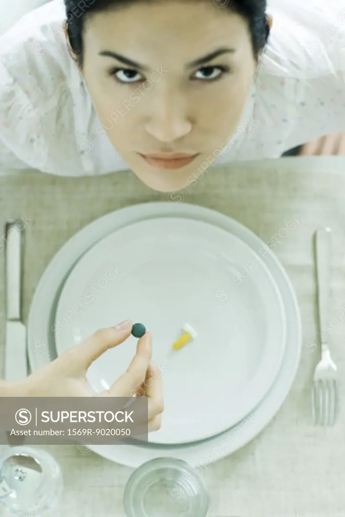 Woman sitting at empty plate holding vitamin, looking up at camera