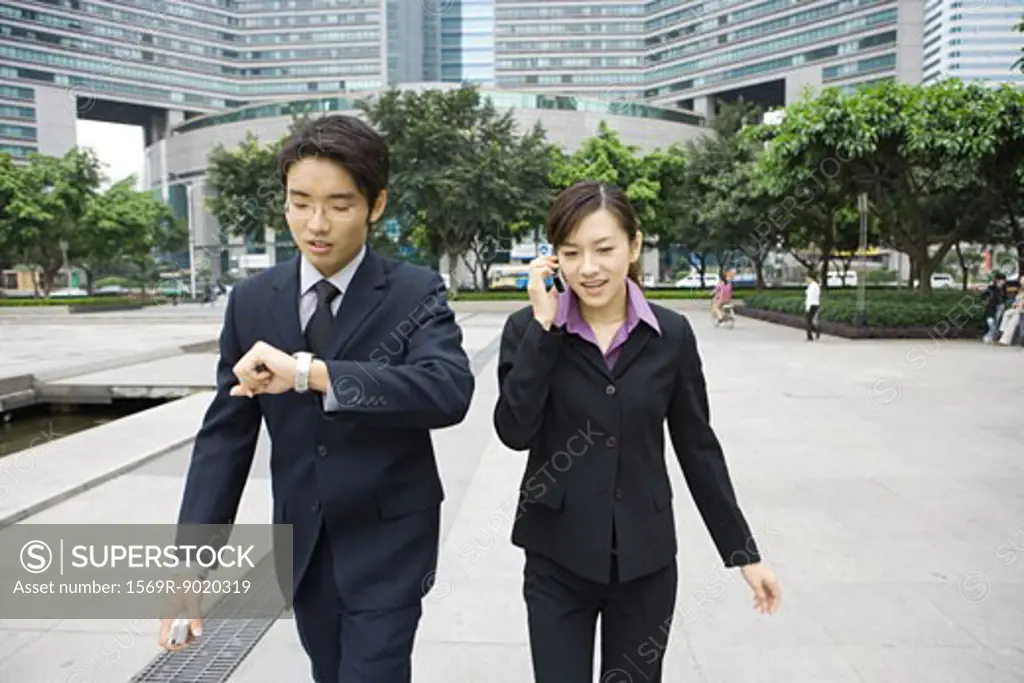 Two young executives walking in office park, one checking watch while other uses cell phone