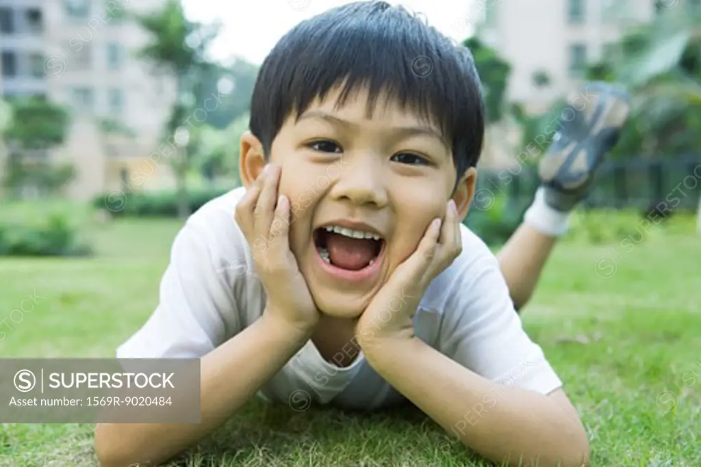 Boy lying on grass, holding head in hands, mouth wide open