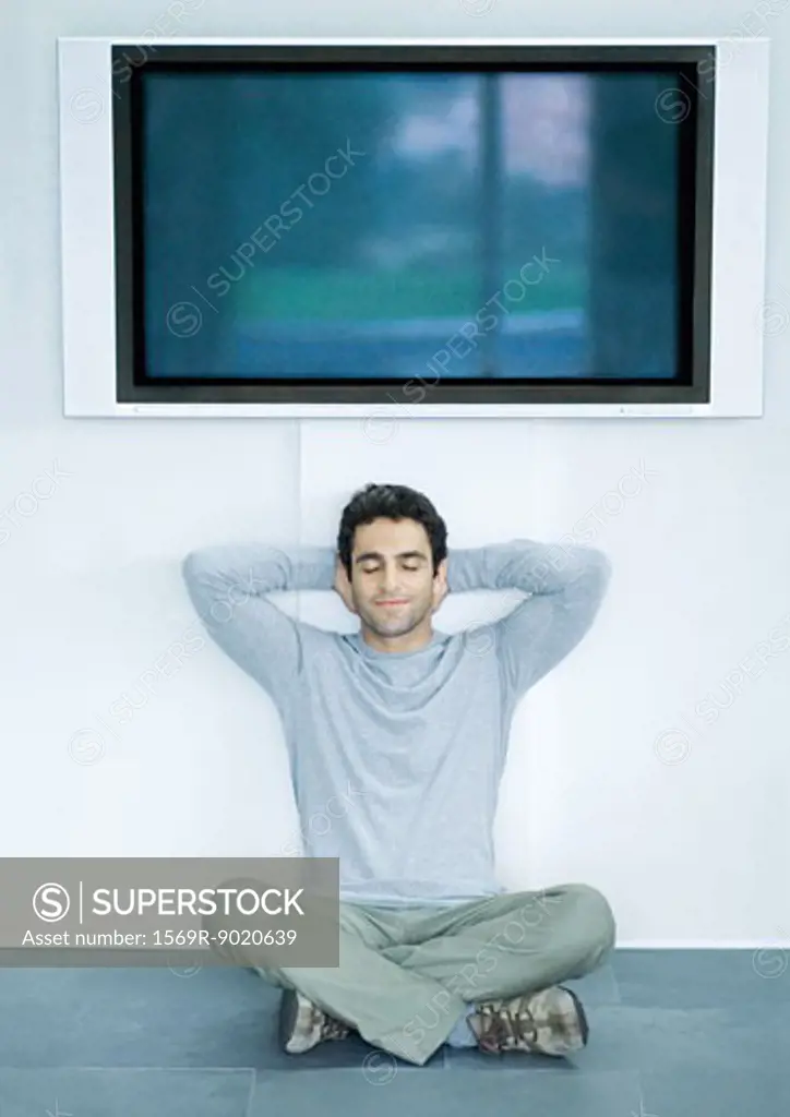 Man sitting on floor under wide screen TV, hands behind head and eyes closed