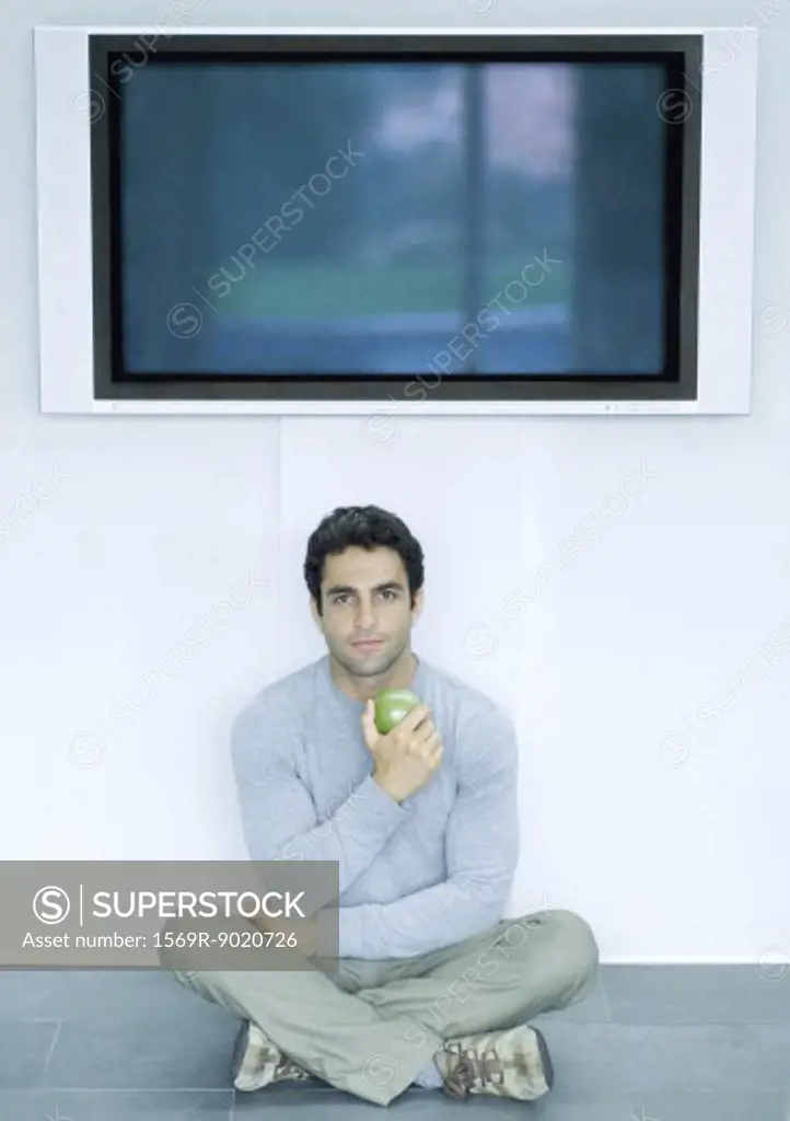 Man sitting on floor under wide screen TV, holding apple