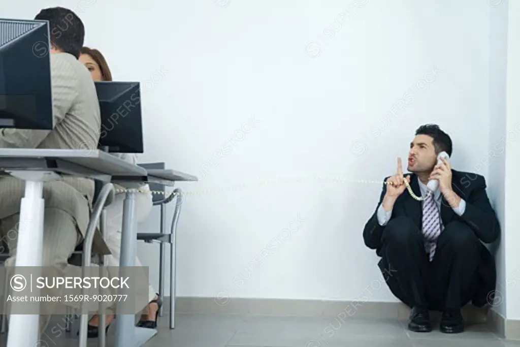 Businessman sitting in corner, holding telephone, looking at colleagues and holding finger in front of lips