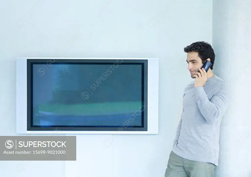 Man leaning against wall using phone, next to widescreen TV