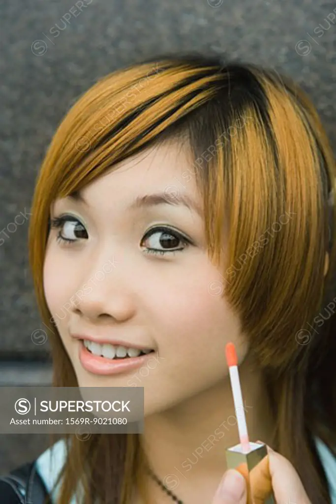 Young woman holding up lip gloss wand, smiling at camera