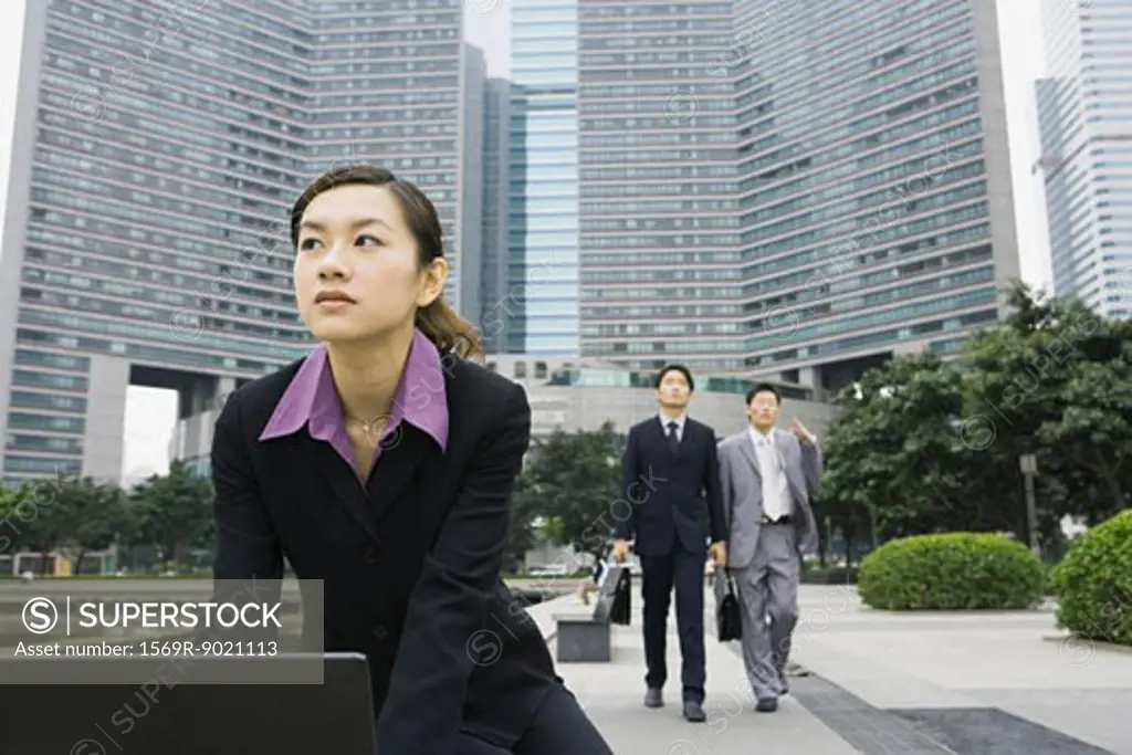 Businesswoman using laptop in office park, two businessmen approaching in background