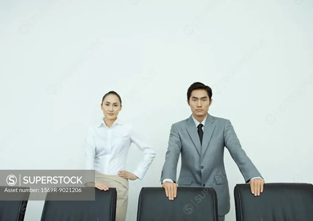 Male and female executives standing with hands on backs of chairs, looking at camera, portrait