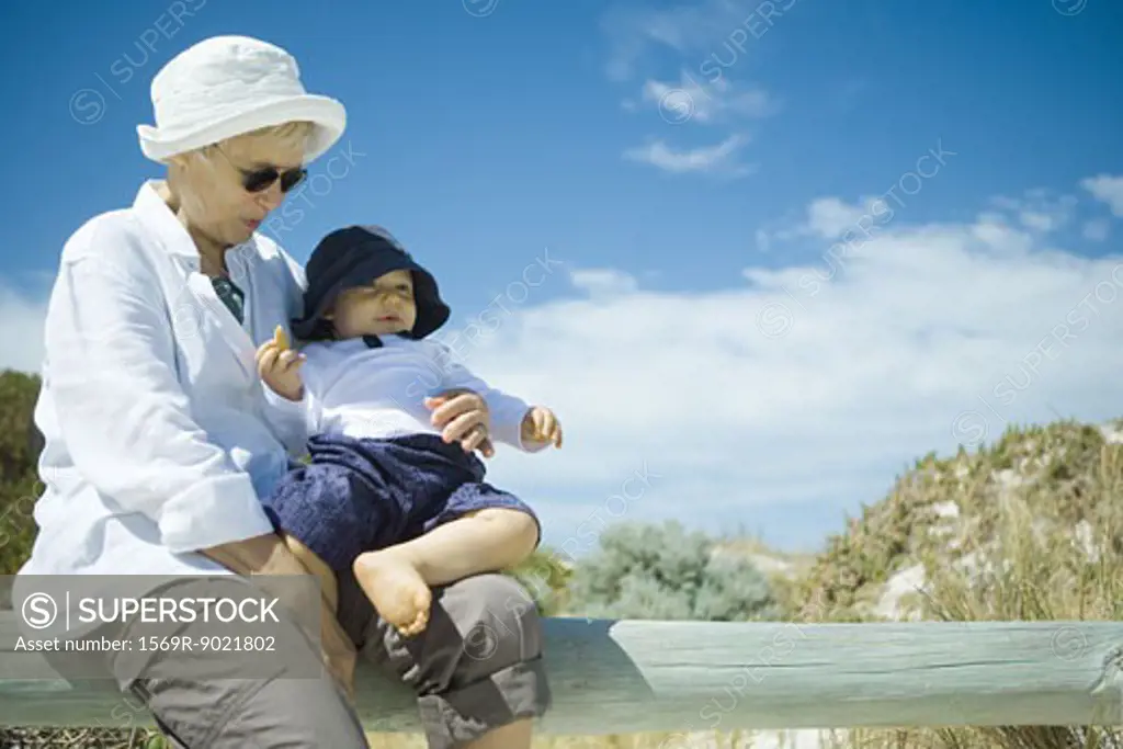 Senior woman sitting on wooden railing, holding baby, dunes in background