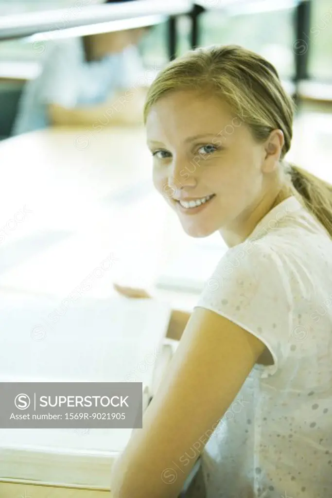 Female college student sitting at table in library, smiling at camera