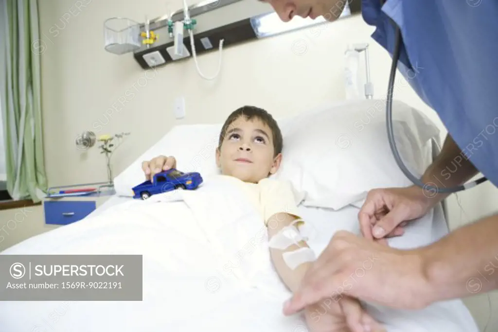 Boy lying in hospital bed holding toy car while intern checks pulse