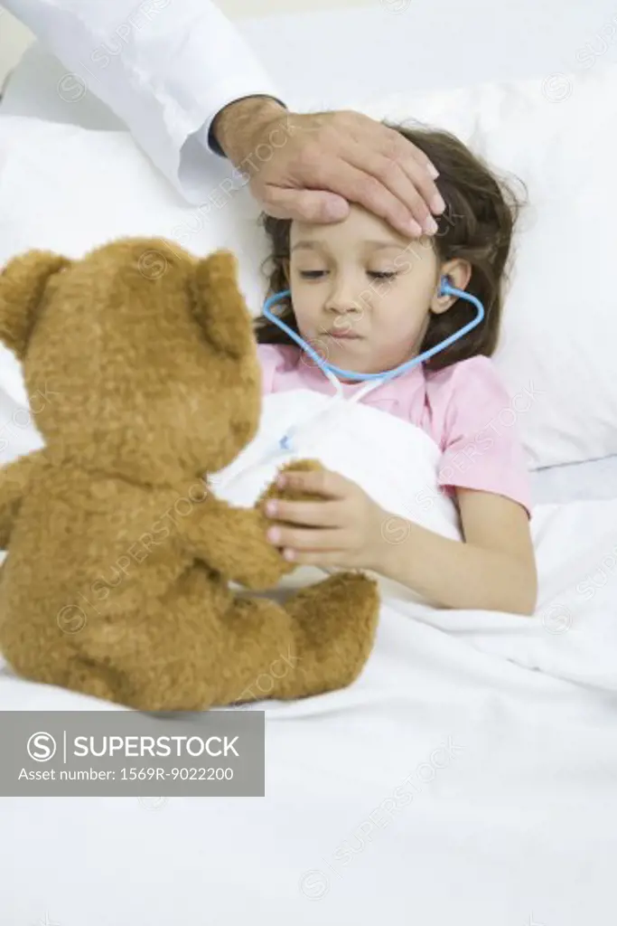 Girl lying in hospital bed, holding teddy bear, doctor's hand on forehead