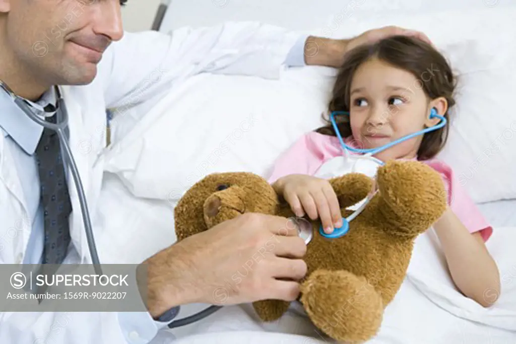 Girl lying in hospital bed, smiling at doctor as both hold stethoscopes to girl's stuffed animal