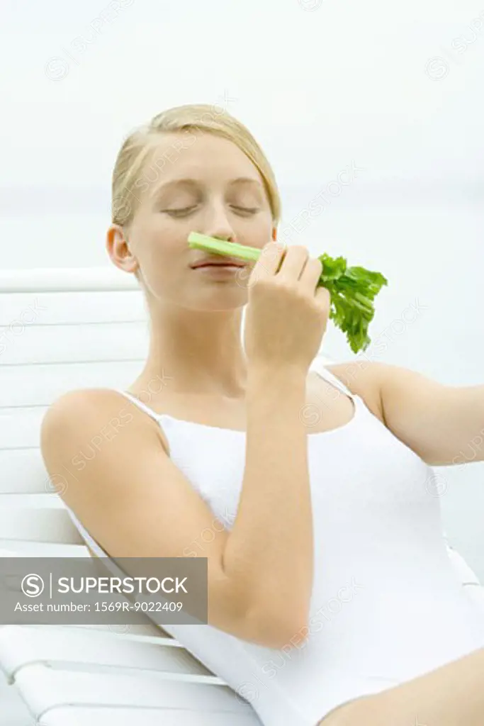 Teenage girl in swimsuit sitting on lounge chair, smelling stalk of celery