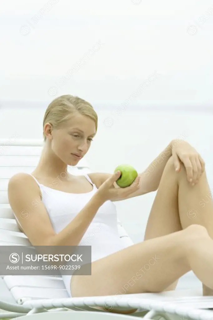 Teenage girl in swimsuit sitting on lounge chair, holding apple