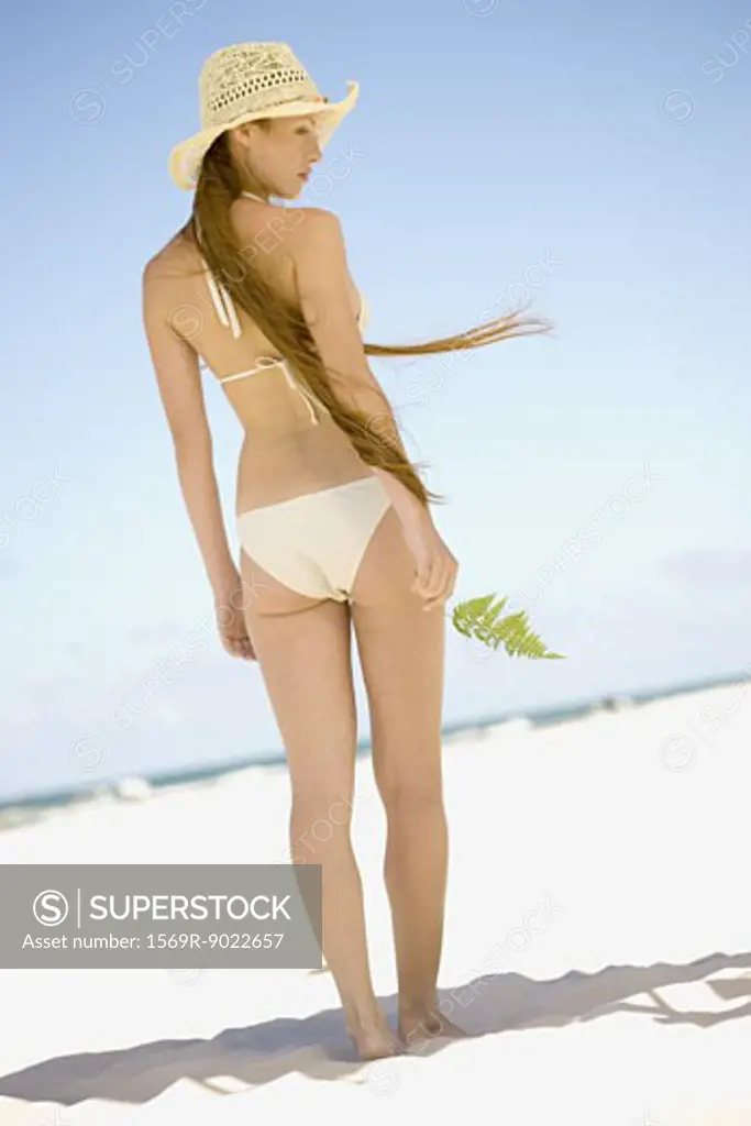 Woman wearing bikini and sunhat on beach, holding leaf in hand, full length, rear view