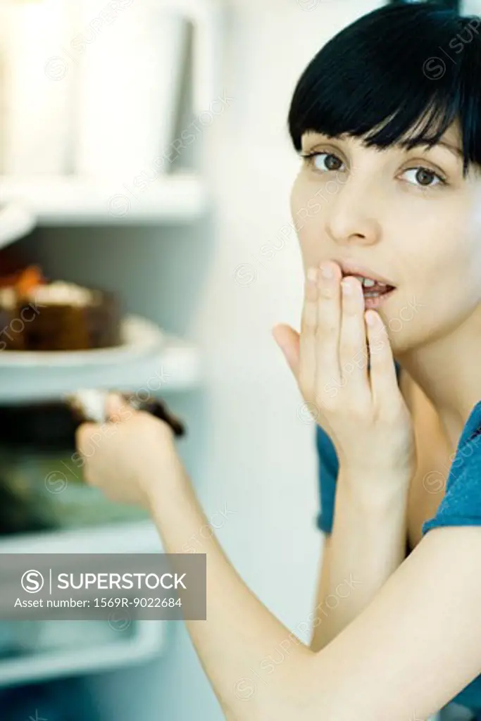 Young woman taking slice of cake from refrigerator, looking at camera, covering mouth