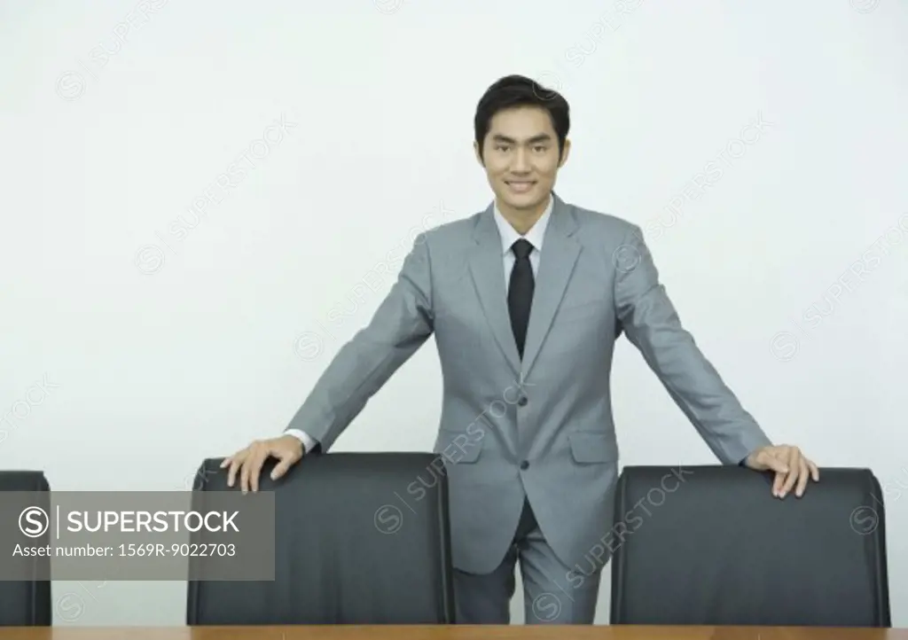 Businessman standing with hands on backs of chairs, smiling at camera, three quarter length