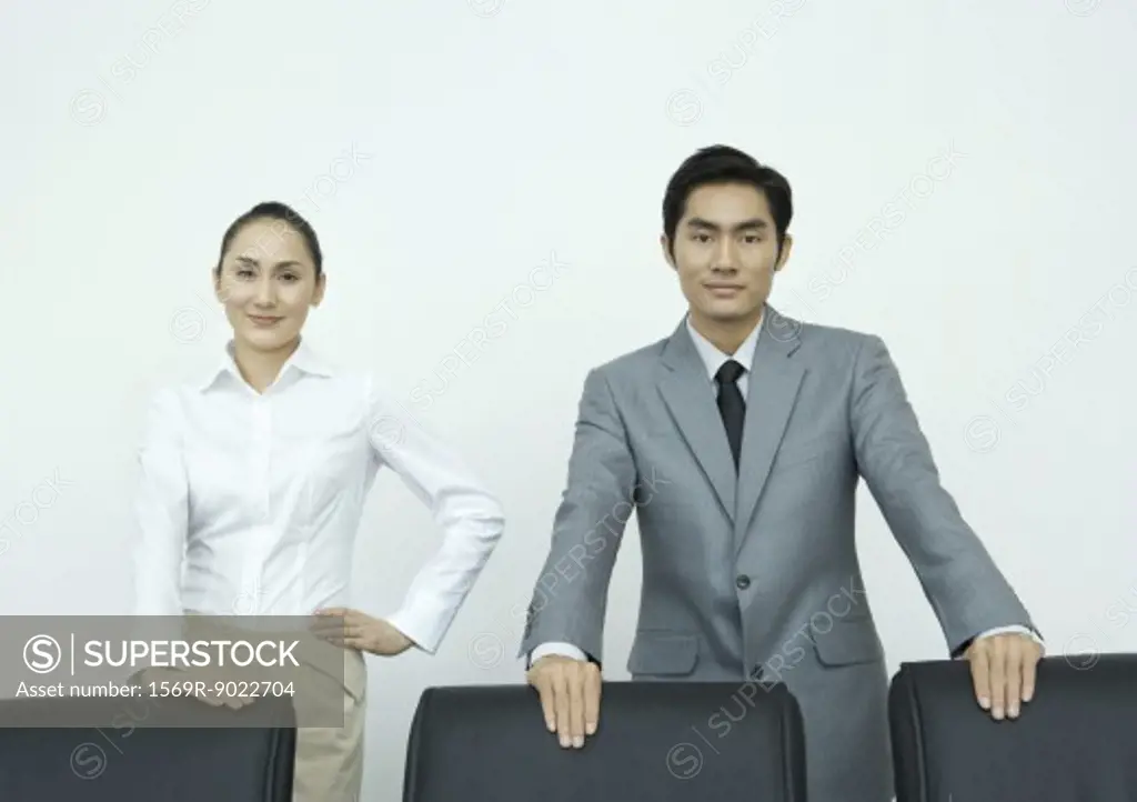 Businessman and businesswoman standing with hands on backs of chairs, smiling at camera