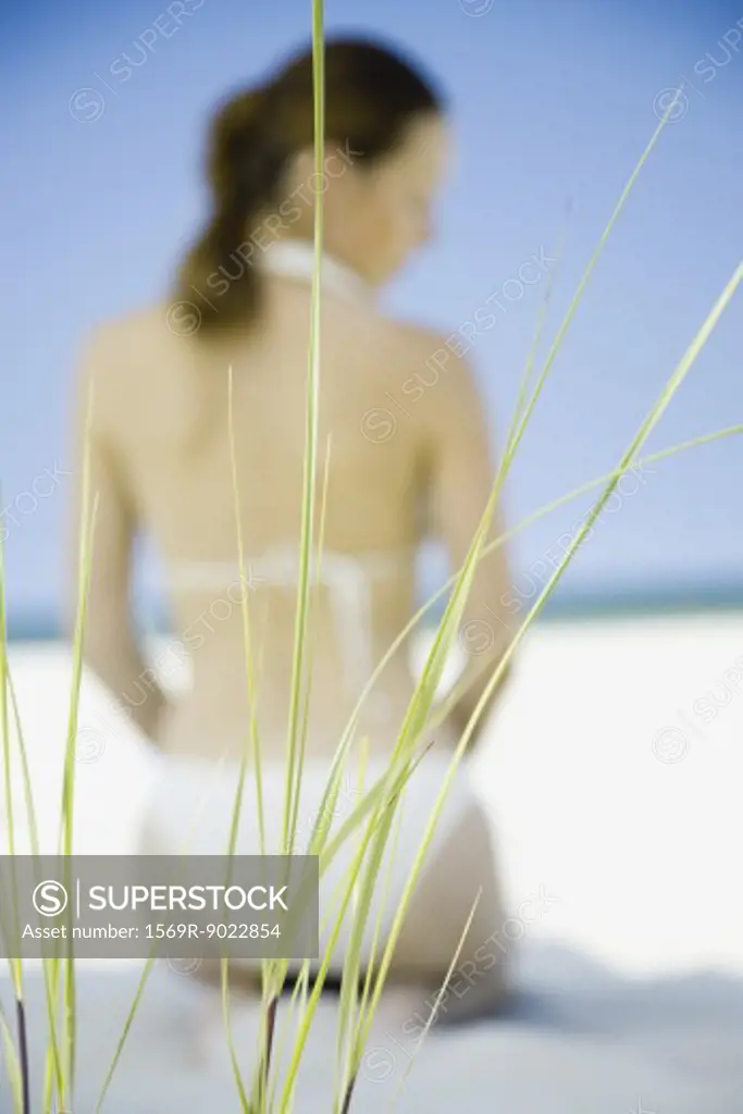 Woman in bikini kneeling on beach, focus on sea oats in foreground