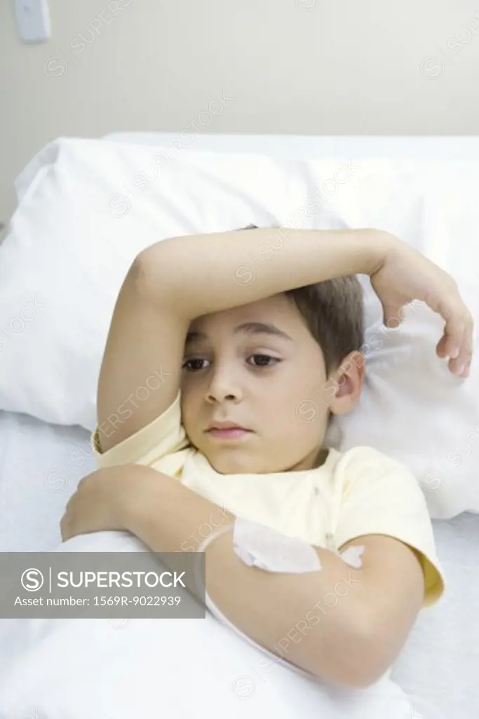 Boy lying in hospital bed with arm across forehead, looking away