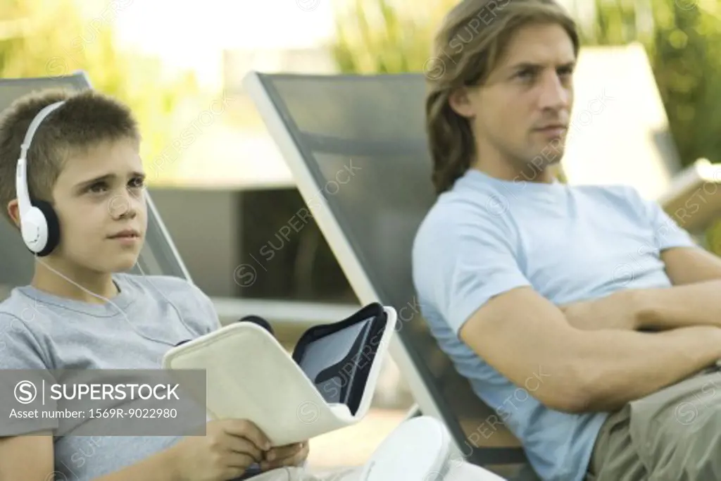 Father and son sitting on lounge chairs together, boy listening to headphones
