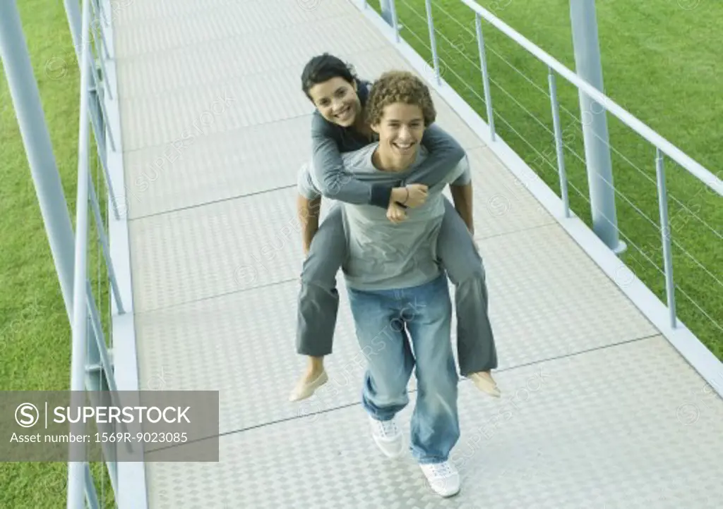Teenage couple, boy carrying girl piggyback on walkway, smiling at camera
