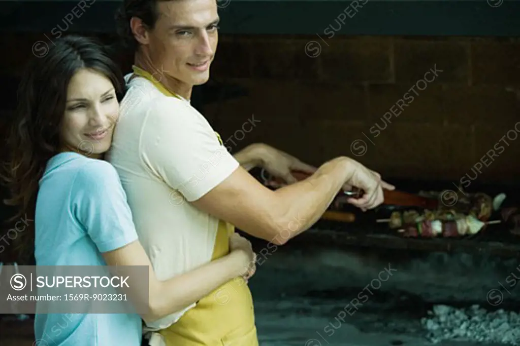 Man tending barbecue, woman hugging man from behind