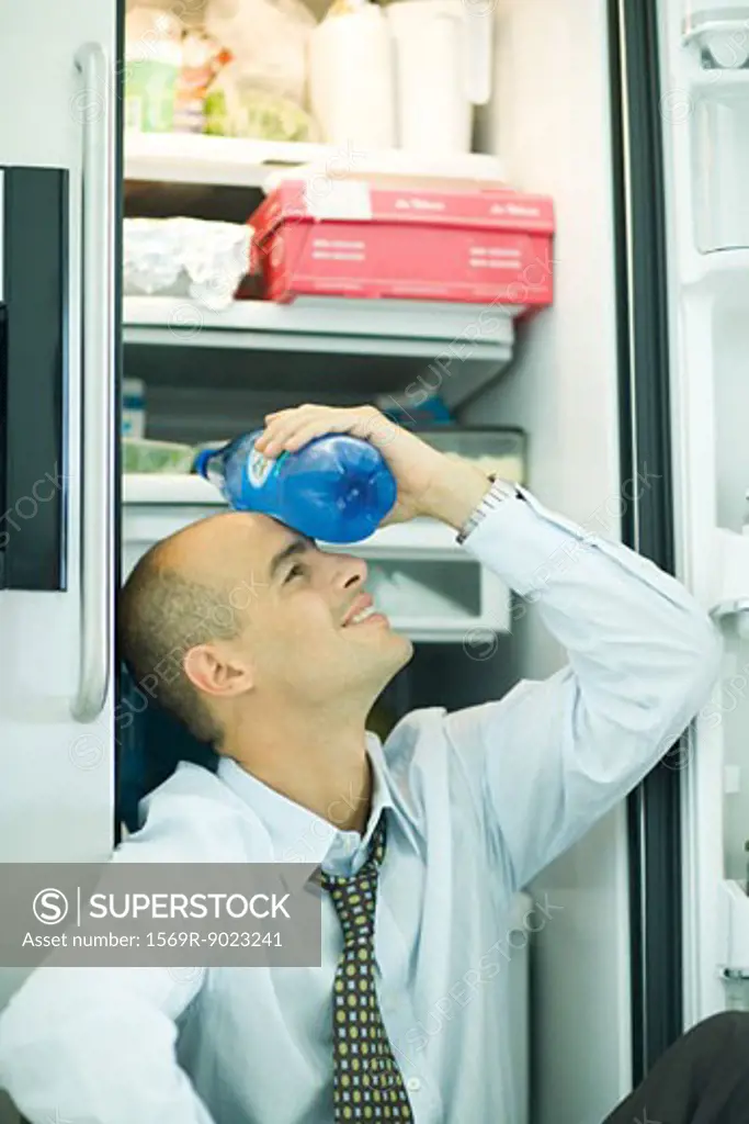 Man sitting in front of open refrigerator, holding bottle of water on forehead