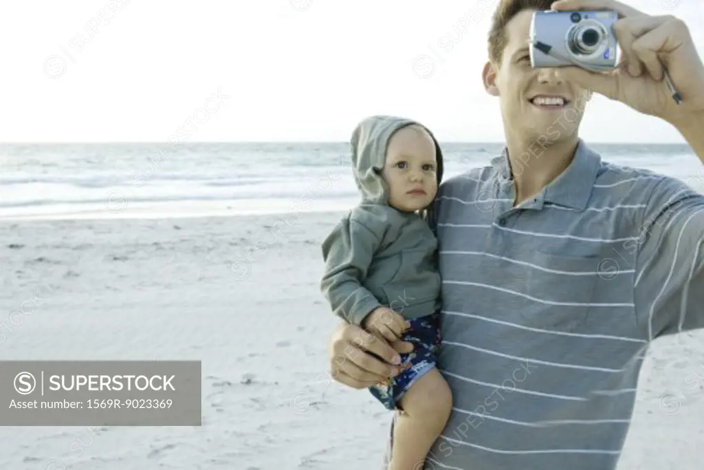 Man holding baby and using camera on beach