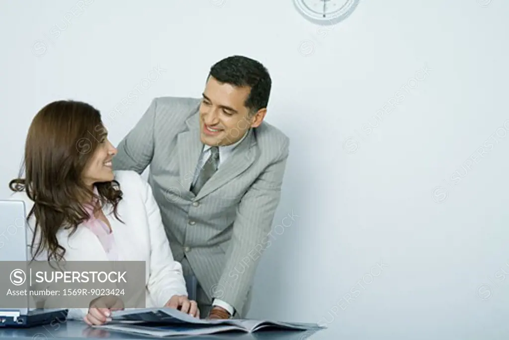 Businessman and businesswoman smiling at each other while woman sits at desk and man looks over her shoulder