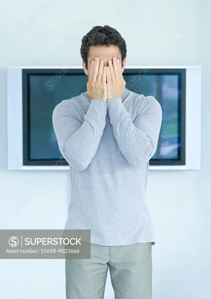 Man standing in front of wide screen TV, hands covering face