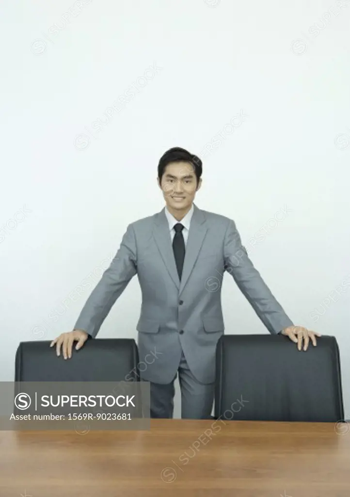 Businessman standing in conference room with hands on backs of chairs, portrait