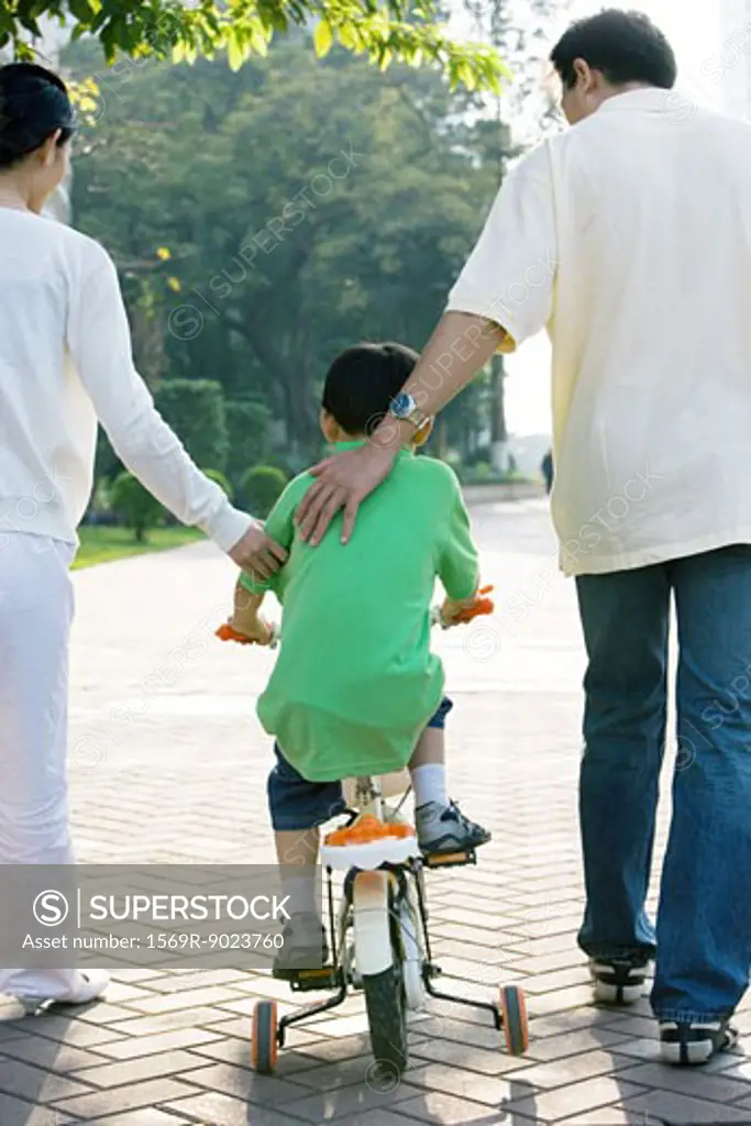 Boy riding bicycle with training wheels, parents on either side, rear view