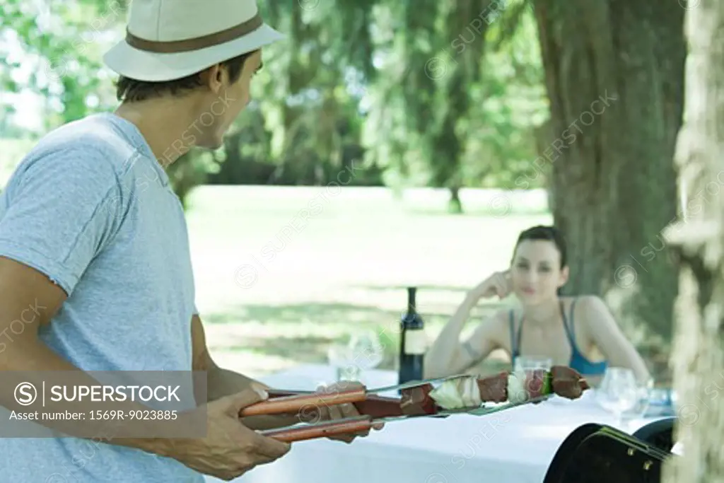 Man holding kebab, turning toward woman sitting at picnic table