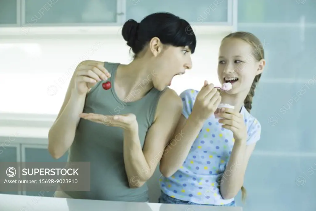 Girl holding ice cream dessert, next to woman holding cherry, woman leaning toward girl with mouth wide open