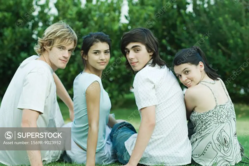 Four young friends sitting on grass, side by side, looking over shoulders at camera