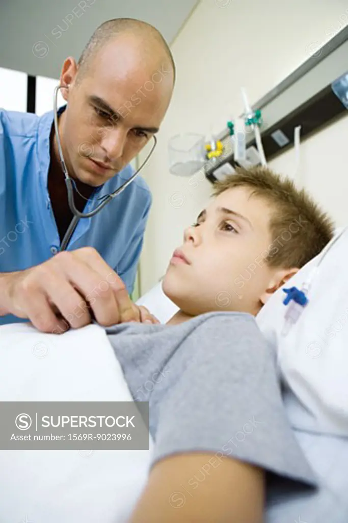 Boy lying in hospital bed, doctor listening to chest with stethoscope