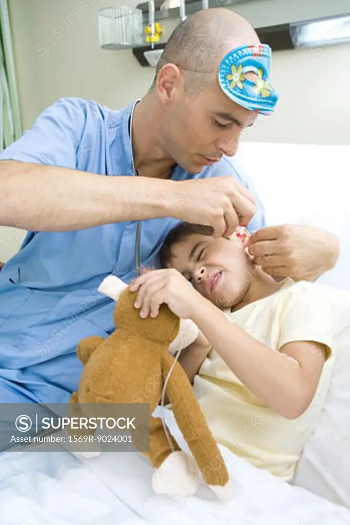 Boy lying in hospital bed, holding stuffed animal while doctor puts drops into boy's ear