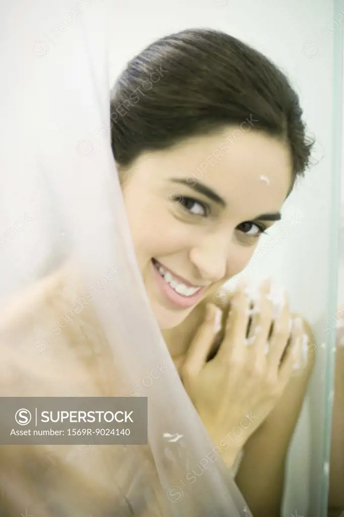 Young woman standing behind shower curtain, smiling at camera