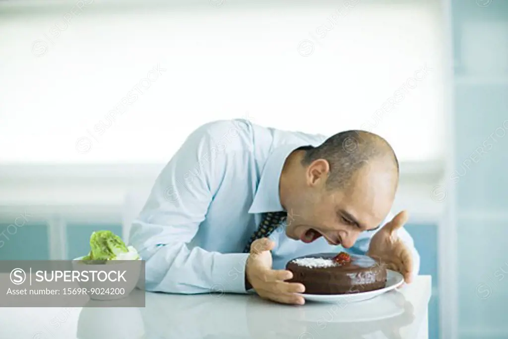 Man sitting at table with chocolate cake and bowl of lettuce, leaning toward cake with mouth wide open
