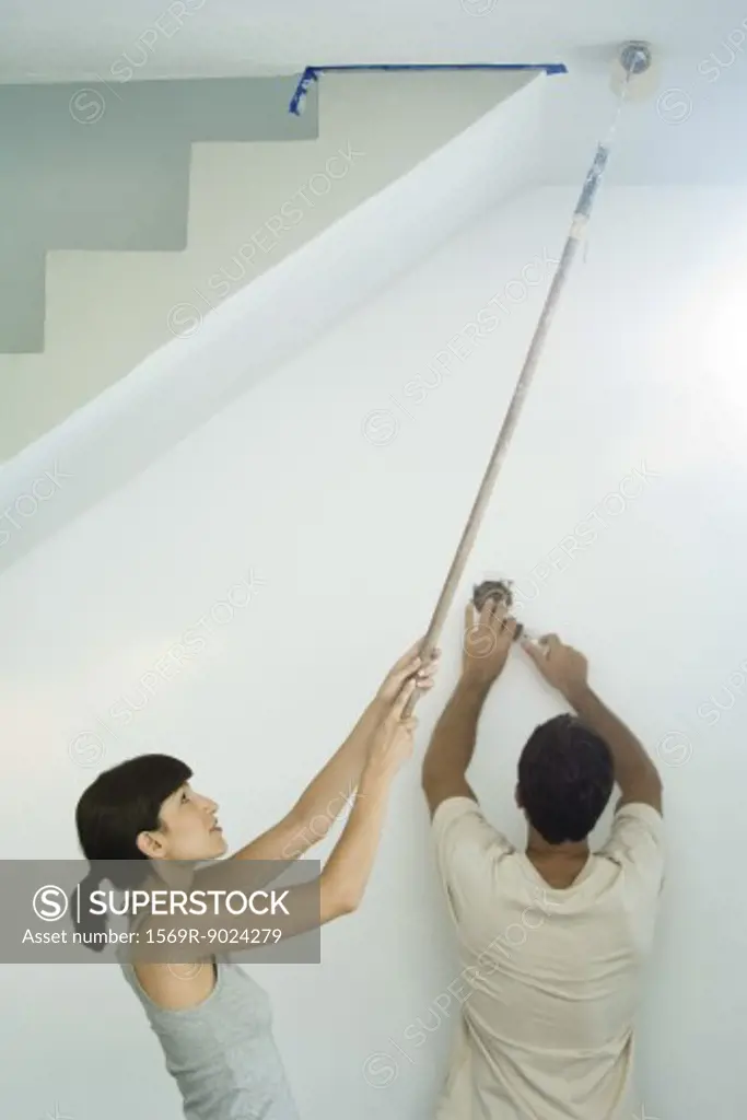 Woman painting ceiling with paint roller while man changes lightbulb