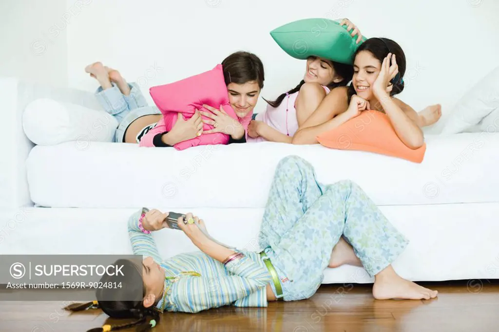 Three young female friends lying down, holding cushions while fourth friend takes photo
