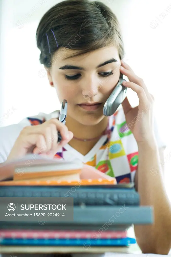 Teen girl with stack of homework, using cell phone and holding pen