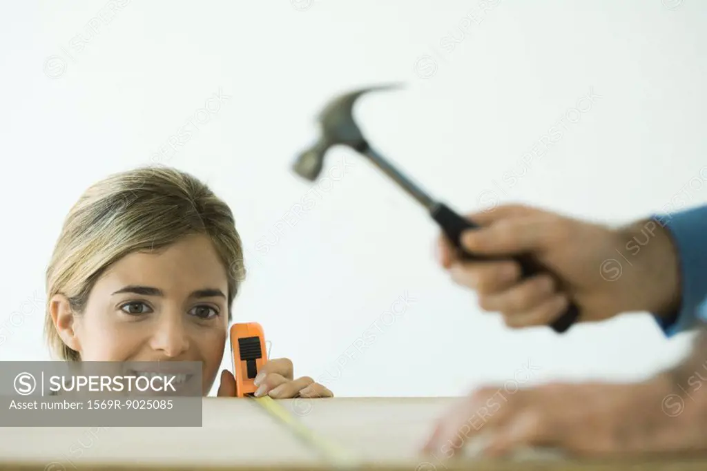 Woman measuring board while man hammers, focus on woman in background
